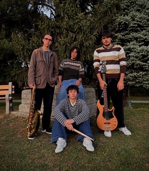 Four band members from The Stack standing together outdoors. One member has a guitar and another has a saxophone.
